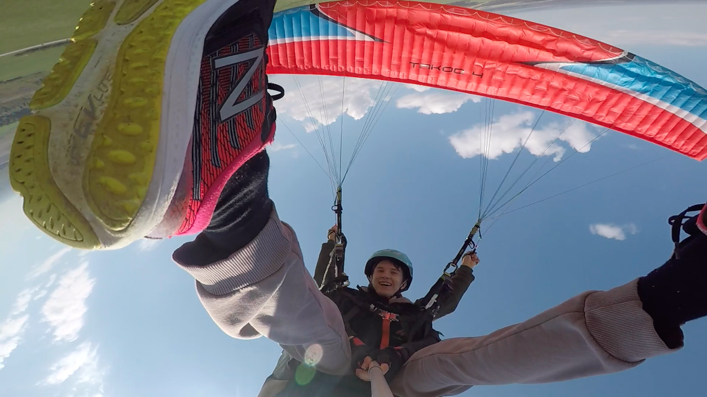 Два парапланериста в тандеме летят над пейзажем, демонстрируя волнение и восторг от полета на параплане.